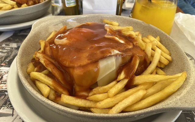 A francesinha sandwich covered in melted cheese and sauce, served with fries