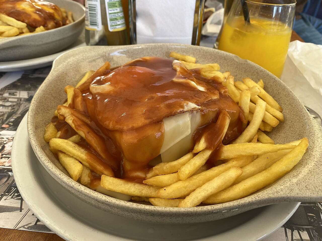 A francesinha sandwich covered in melted cheese and sauce, served with fries